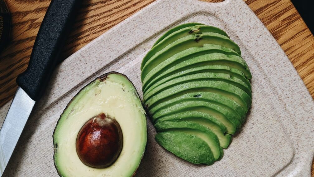 an avocado that has been cut in half resting on a cutting board, one half has been peeled, sliced horizontally and fanned out