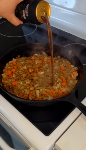 Guinness stout being poured into a heavy cast iron pan. The beer is flowing out of the can onto fried onions, carrots, lentils, and garlic.