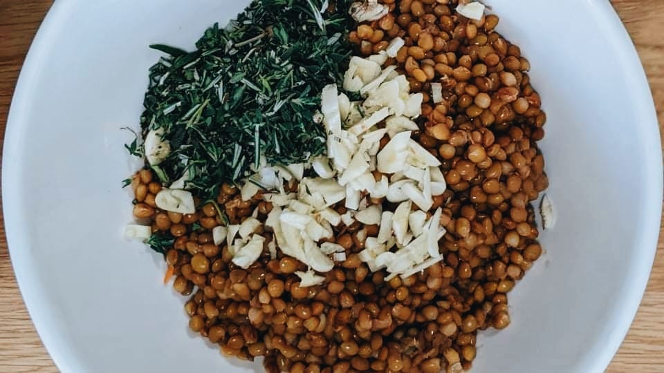 A bowl containing chopped rosemary, thyme, garlic, and rinsed lentils.