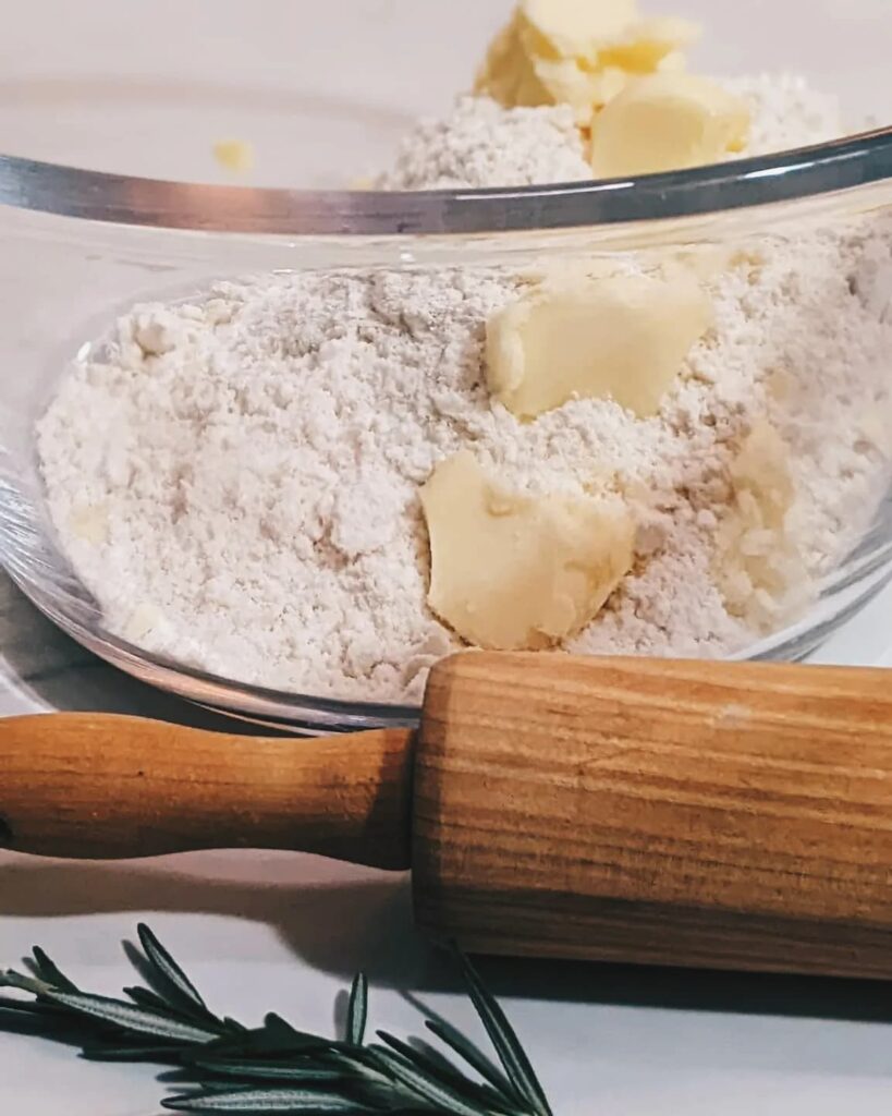 A medium sized mixing bowl containing flour, salt and butter. Beside the bowl is a rolling pin, and a sprig of rosemary. This will become a pie/galette dough.